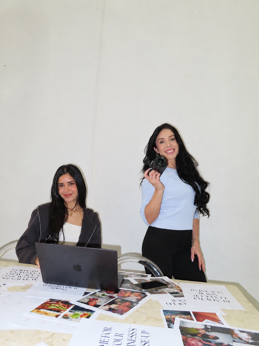 Two women in a workspace setting, with one seated at a table using a laptop and the other standing holding a smartphone. Papers and printed photos are spread across the table against a plain off-white wall.